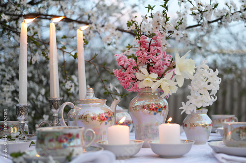 Romantic candlelit dinner in the garden. Table setting in light colors. Bouquet in a vase of pink hyacinths and white daffodils. Blossoming trees in the background.

