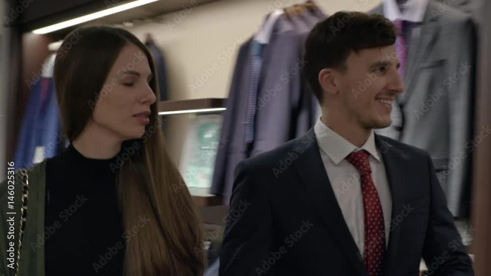 Tracking shot of young man in suit and his female friend walking in menswear clothing store and choosing jacket Tracking shot of young man in suit and his female friend walking in menswear clothing store and choosing jacket