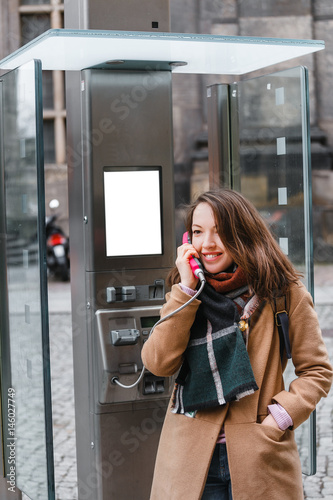 Portrait of happy and smiling tourist woman talking in the modern payphone on the city street, telephone connection during a trip concept