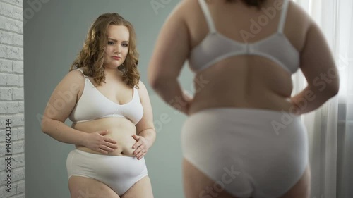 Obese lady in underwear looking at reflection in mirror, ashamed of her fat body