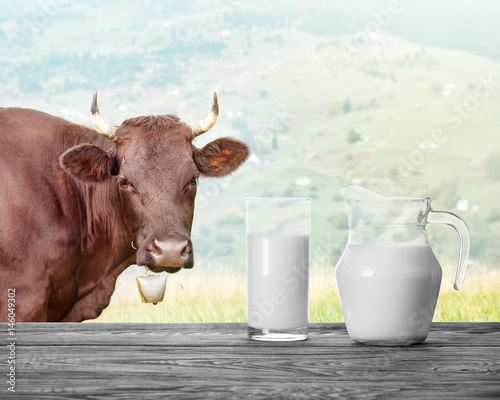 Jug and a glass of milk against background of landscape with cow