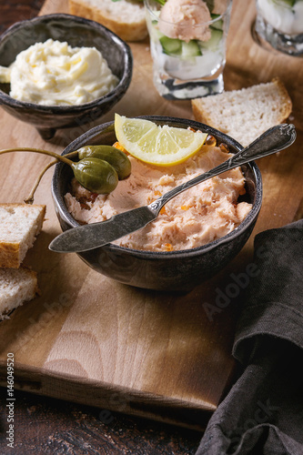 Black bowl of salmon pate with red caviar served with butter, sliced bread, capers, vintage knife, verrines and herbs on wooden serving board, textile linen over brown texture background. Close up