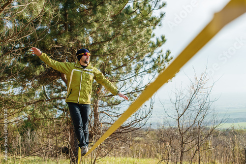 A man, aged with a beard and wearing sunglasses, balances on a slackline in the open air between two trees at sunset