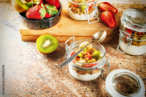 Healthy breakfast. Diet. Overnight oatmeal in a can, muesli. Yogurt with homemade granola and organic fruits - kiwi, banana, strawberry. On the stone table. With ingredients and spoons. 