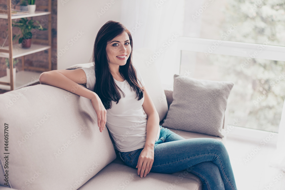 Young pretty brunette girl is relaxing at home sitting on nice sofa. She is wearing casual clothes, smiling and looking straight Young pretty brunette girl is relaxing at home sitting on nice sofa. She is wearing casual clothes, smiling and looking straight