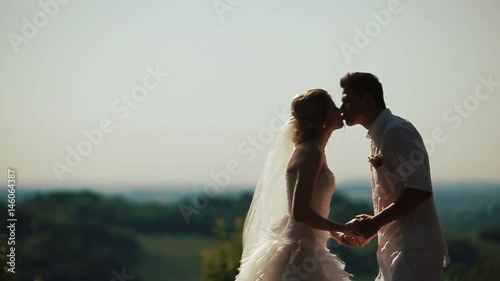 Gorgeous young wedding couple in summer dresses kisses before green landscape