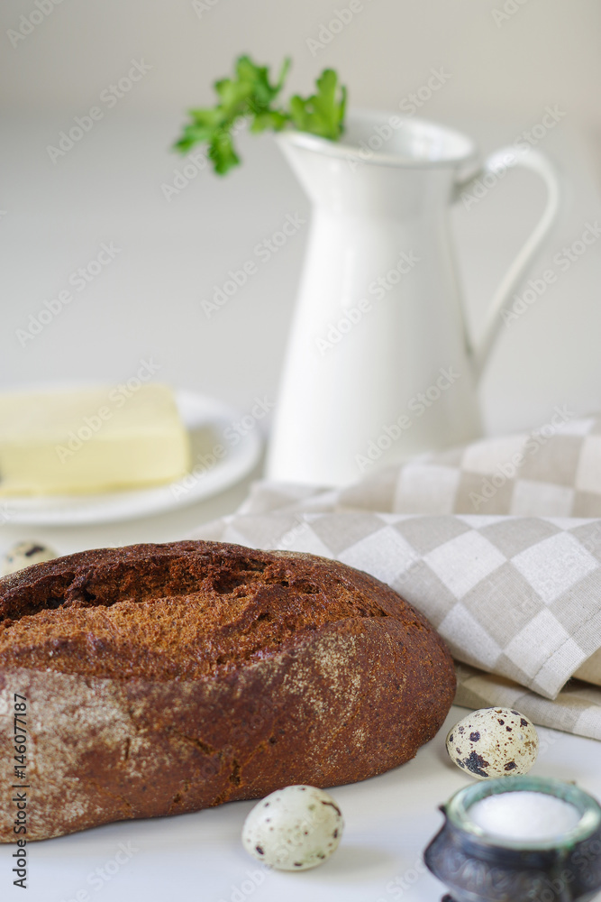 Loaf of rye bread on linen napkin with  white jug, salt, quail eggs and butter on background