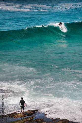 Photography surfer stand on the rocks watching a buddy  surfing a wave on the coast between Bondi and Bronte, Sydney, Australia