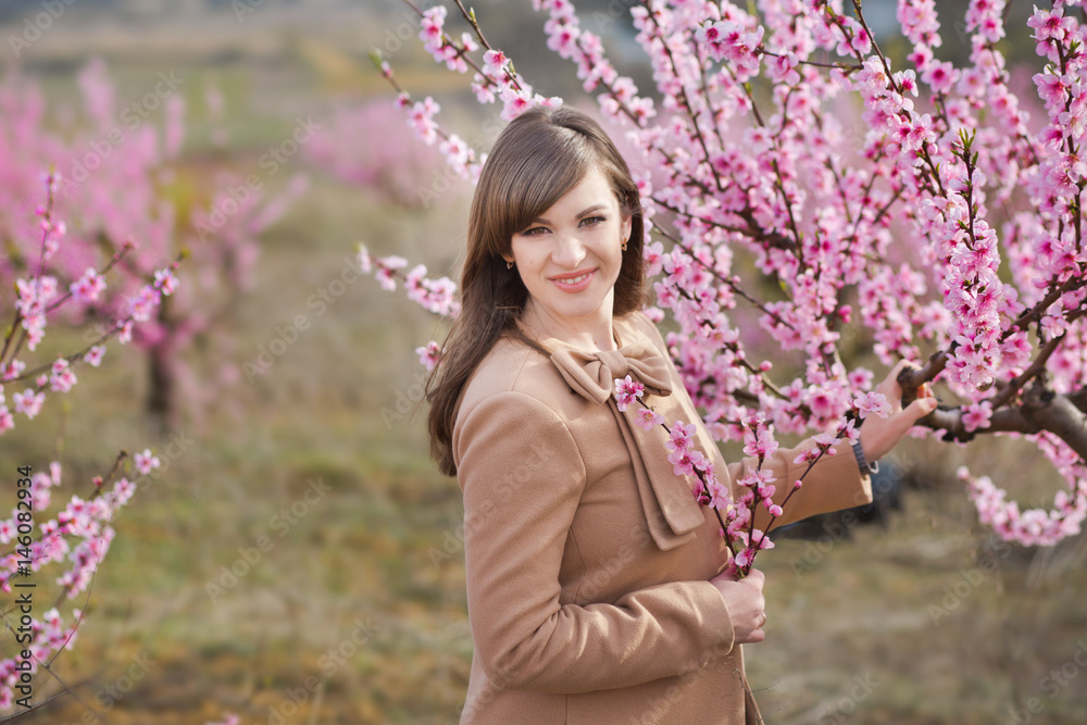 Cute beautiful stylish dressed brunette girl with mother mom standing on a field of spring young peach tree with pink flowers.Lady dressed in rain clothes and blue hat rubber boots spring season. Cute beautiful stylish dressed brunette girl with mother mom standing on a field of spring young peach tree with pink flowers.Lady dressed in rain clothes and blue hat rubber boots spring season.