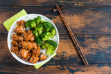 Chicken lacquered with a sweet soy teriyaki sauce in a white bowl. Garnished with rice and broccoli. Chopsticks, brown wooden table, top view.
