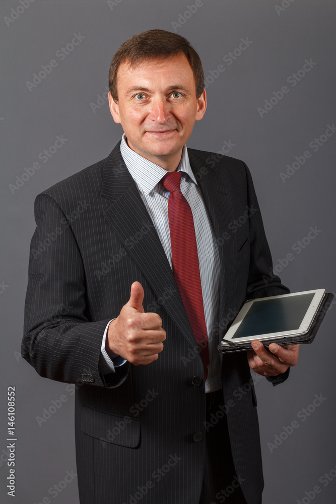 Confident mature businessman standing in front of a grey background holding a notebook Confident mature businessman standing in front of a grey background holding a notebook