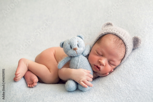 Newborn boy in a naked hat lies on a light blanket with a blue knitted bear