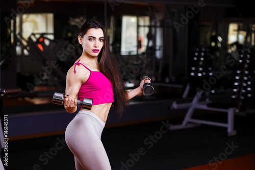 Young attractive muscular smiling fitness woman doing exercise with dumbbells in gym