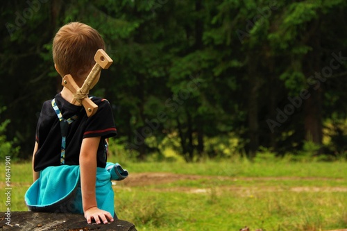 Boy warrior with a wooden toy sword looking at the forest, back view