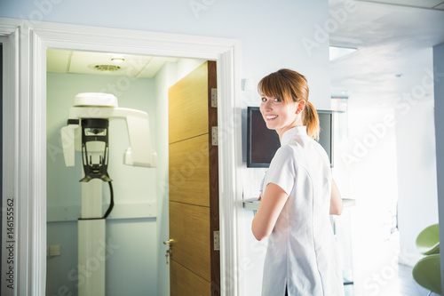 Happy smiling doctor or assistent of stomatology clinic opened the door of cabinet with orthopantomograph, indoors