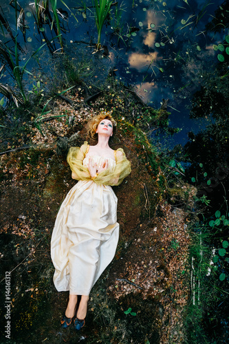 Slika na platnu Young Russian witch faery bloat girl fish queen in fairy dress lying on swamp shore with sky, plants reflected in water