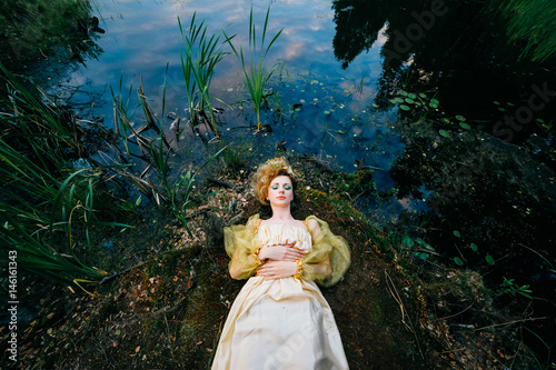 Young Russian witch faery bloat girl fish queen in fairy dress lying on swamp shore with sky, plants reflected in water. Colorful, magic and classical literature tale idea. Fantasy concept. Sorcery. photo