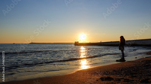 A girl is standing by the sea at sunset