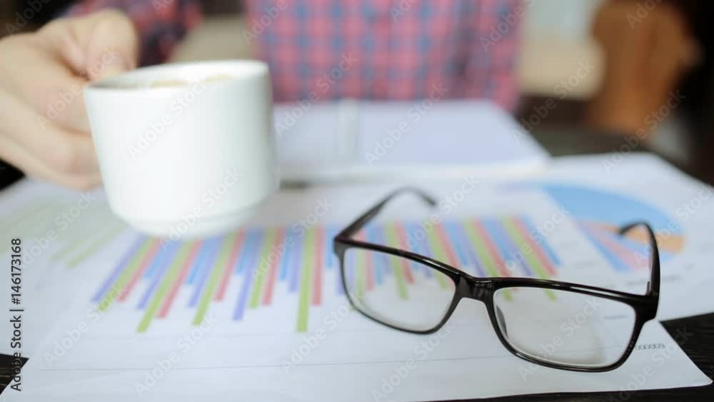 Close up hand of business man holding cup of coffee with paper graph chart on wooden desk Close up hand of business man holding cup of coffee with paper graph chart on wooden desk