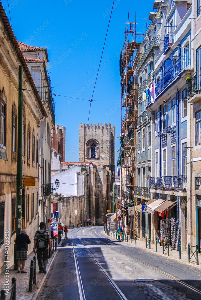 Steep narrow street of Lisbon with cable car rails in the middle and fort tower in the background