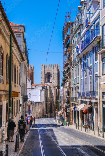Fototapeta Naklejka Na Ścianę i Meble -  Steep narrow street of Lisbon with cable car rails in the middle and fort tower in the background
