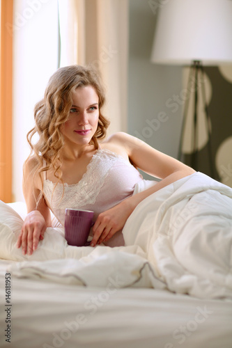 Young woman drinking cup of coffee or tea while lying in bed