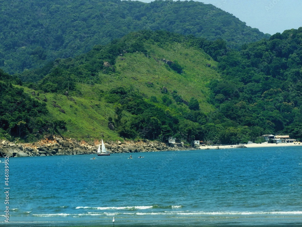 Praia de águas azuis vista do mar com barco ao longe e  atrás montanha com floresta e vegetação de vários tons de verde 