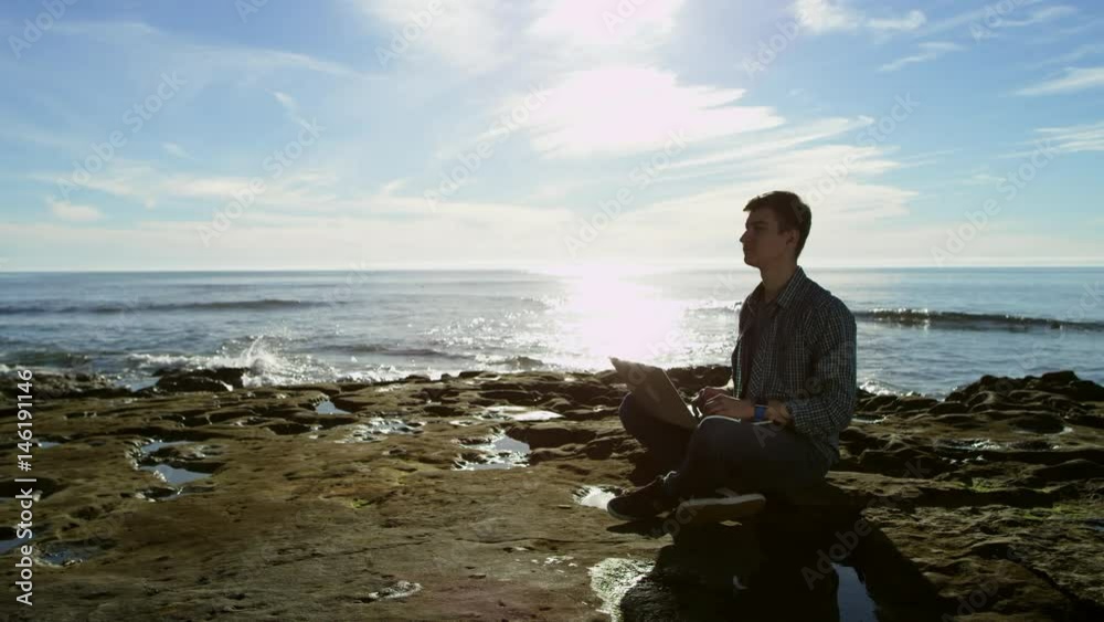 Attractive youthful male is chatting with his friends over the Internet while sitting on a beach Attractive youthful male is chatting with his friends over the Internet while sitting on a beach