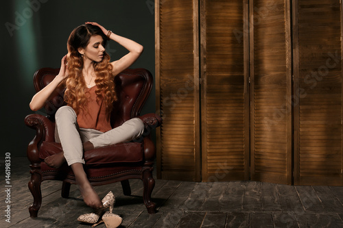 Fashion model in fashion clothing sitting in brown leather armchair posing in the studio.
