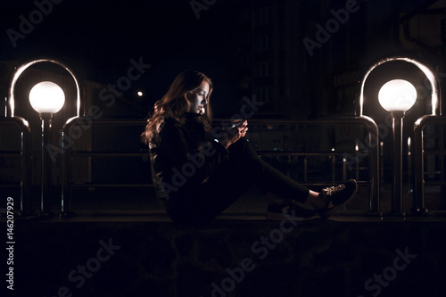 Caucasian teenage girl sitting on bench at night texting on cell phone