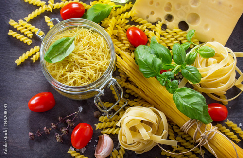 Various kinds of pasta ingredients with basil, cherry tomato, garlic, pepper and cheese on wooden kitchen table. The concept of Italian food.