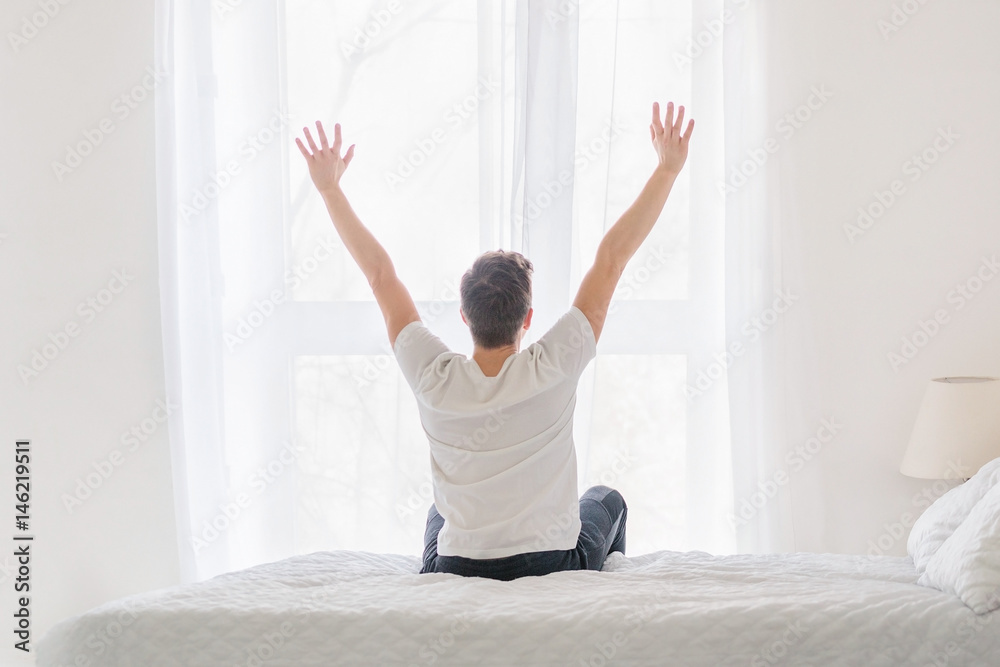 Young man sitting on bed with hands raised up in the morning. Ready for new day. Rear view Young man sitting on bed with hands raised up in the morning. Ready for new day. Rear view
