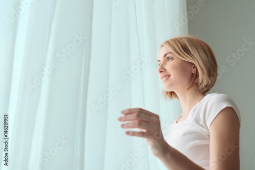Happy smiling woman looking out through the curtain at window at home