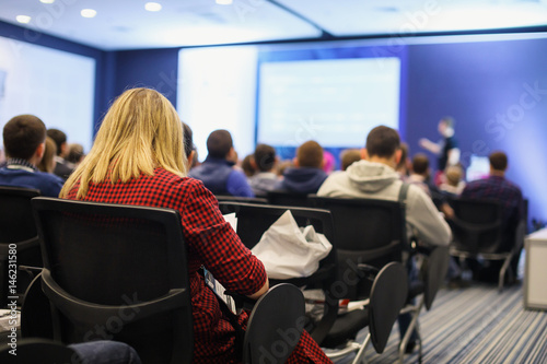 Business and entrepreneurship symposium. Speaker giving a talk at business meeting. Audience in the conference hall. Rear view of unrecognized participant in audience.