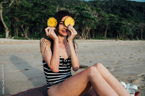 Beautiful young woman tourist In a striped swimsuit holding two ripe pineapple against her eyes. photo