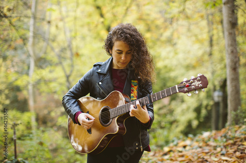 Beautiful young woman playing guitar on forest, fashion lifestyle. Girl wearing black jacket.