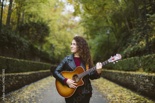 Beautiful young woman playing guitar on forest, fashion lifestyle. Girl wearing black jacket.