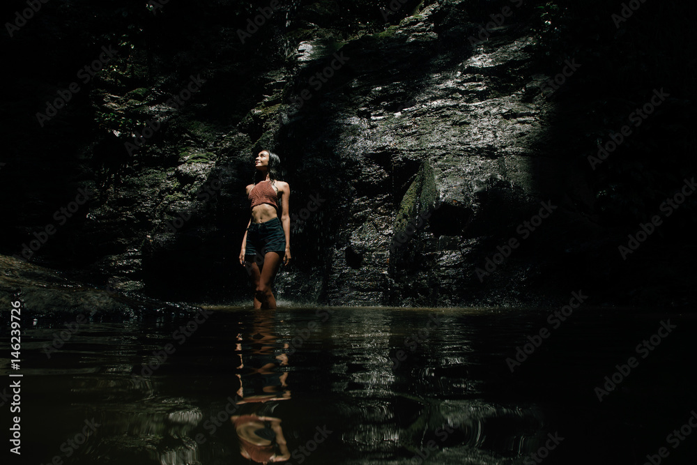 Fototapeta premium beautiful wet girl in shorts and a knitted blouse against a waterfall background. Dark background.