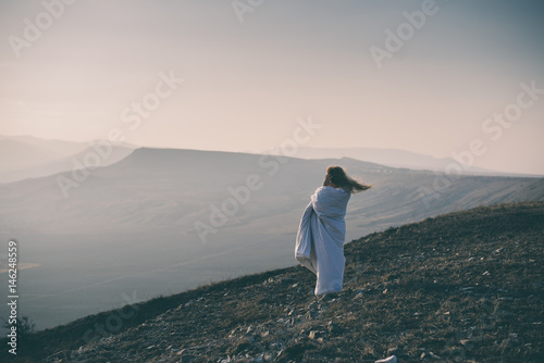 Lonely sad and crying young woman sitting wrapped in blanket on cliff of mountain. Toned image