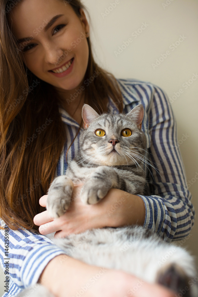Loving girl huging her cat at home near the window. Cheerful girl holding her interested pet paws. Girl loves her cat. Friends expressing feelings