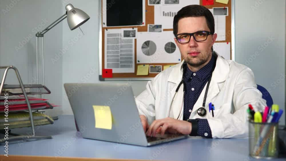 A doctor at a private clinic who wore a white robe and a stethoscope around his neck sits at the desk, his hands on the laptop keyboard. A man with glasses looks at the camera A doctor at a private clinic who wore a white robe and a stethoscope around his neck sits at the desk, his hands on the laptop keyboard. A man with glasses looks at the camera
