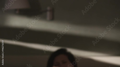 Young pretty drowsy caucasian brunette woman sits on bed and caress hair. Beam of light falls on her face in early morning, close up slow motion, warm tone