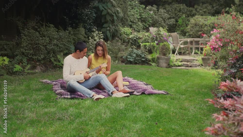 A young couple on a picnic blanket in a backyard, playing a ukulele A young couple on a picnic blanket in a backyard, playing a ukulele