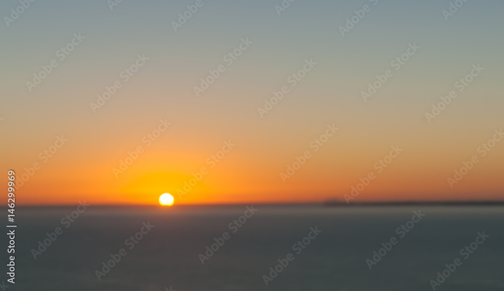 Abstract effect defocused background image Orange glow of sunrise sitting on horizon over blue sea panorama.