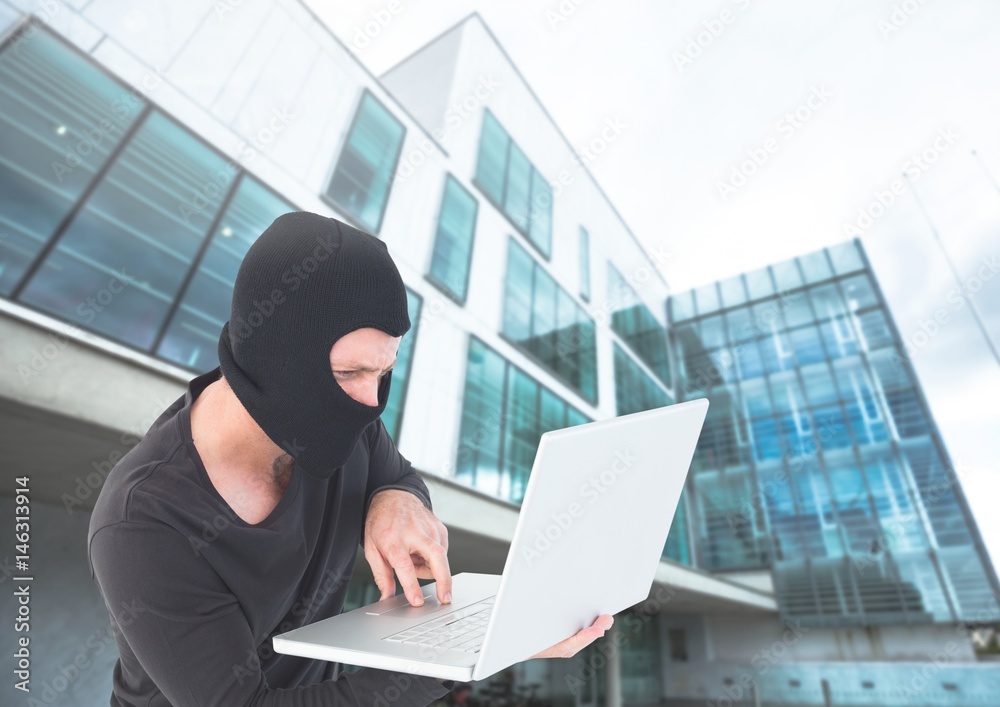 Criminal Man in balaclava on laptop in front of glass building Criminal Man in balaclava on laptop in front of glass building