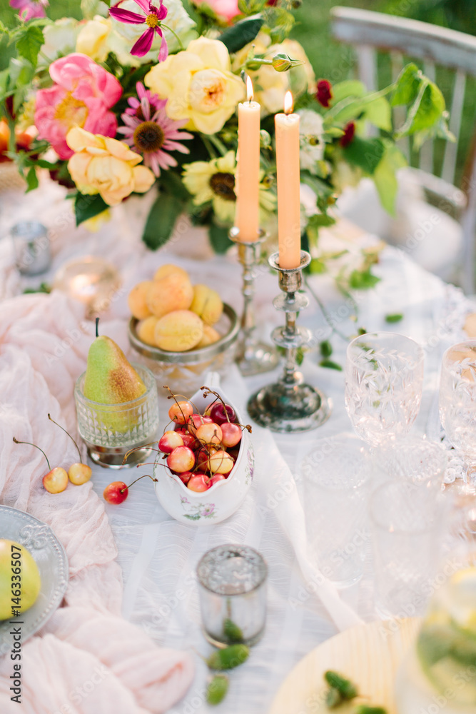 picnic, food, summer, holiday concept - close-up on part of decorated table with beautiful bouquet of roses and pink peonies, burning candles in candlesticks, fruits, glassware, decor of acorns