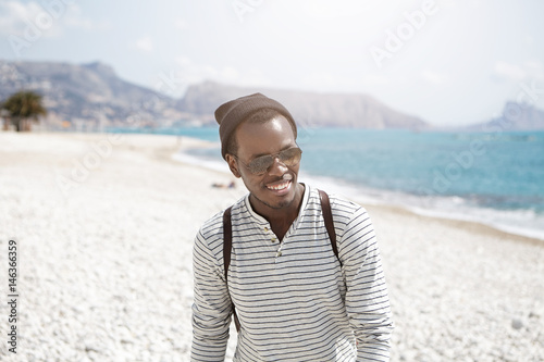 Headshot of laughing young handsome dark-skinned backpacker in mirrored lens sunglasses and hat feeling happy while walking down beach during summer weekend against background of blue sea and rocks