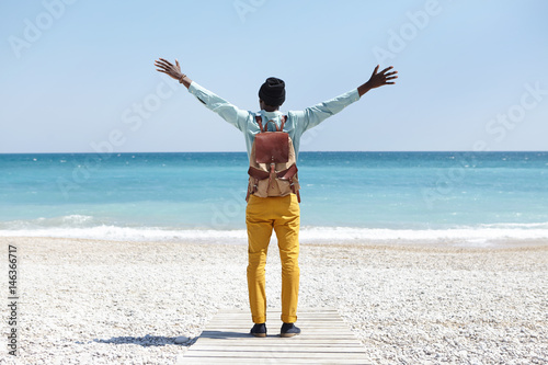 Back view of unrecognizable dark-skinned European man standing on boardwalk on tropical beach feeling happy and free while seeing ocean for the first time during summer trip, keeping arms wide open