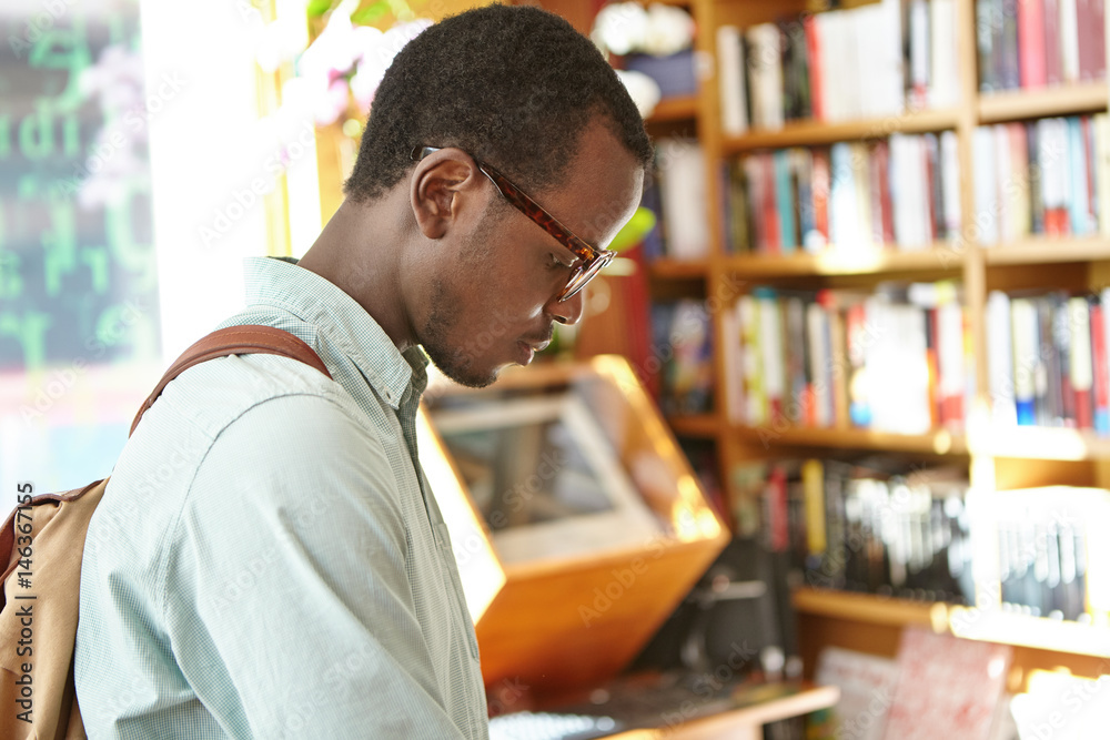 Fototapeta premium Candid shot of concentrated black European male student with backpack working on research in college library. Stylish dark-skinned man looking for phrase book in bookshop before vacations abroad