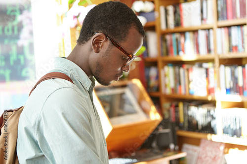 Candid shot of concentrated black European male student with backpack working on research in college library. Stylish dark-skinned man looking for phrase book in bookshop before vacations abroad photo